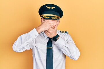 Handsome middle age man with grey hair wearing airplane pilot uniform covering eyes and mouth with hands, surprised and shocked. hiding emotion