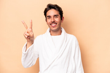 Young caucasian man wearing a bathrobe isolated on beige background joyful and carefree showing a peace symbol with fingers.