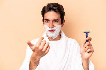 Young caucasian man shaving his beard isolated on beige background pointing with finger at you as if inviting come closer.