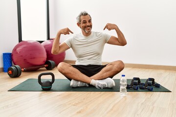 Middle age hispanic man sitting on training mat at the gym looking confident with smile on face, pointing oneself with fingers proud and happy.