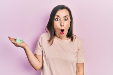 Middle age hispanic woman holding cosmetic moisturizer facial cream scared and amazed with open...