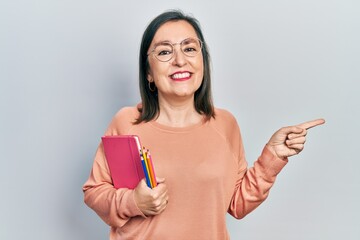 Middle age hispanic woman holding book and color pencils smiling happy pointing with hand and finger to the side