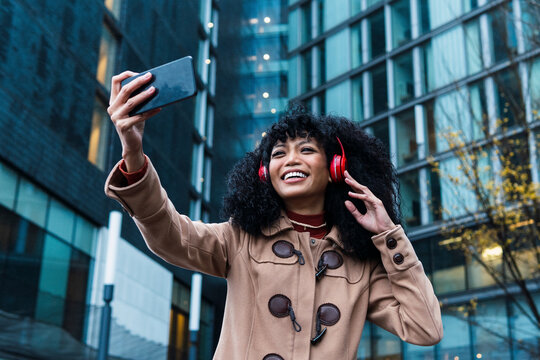 Happy young woman wearing wireless headphones taking selfie through mobile phone in city