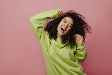 Happy european young woman expresses positive emotions with eyes closed while laughing indoors. Beauty with curly hair keeps hands at face level. People sincere emotions lifestyle concept.