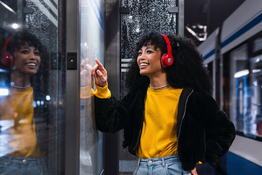 Cheerful young woman wearing wireless headphones searching on map at tram station at night