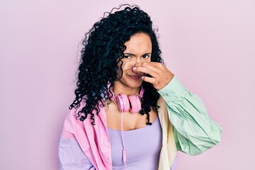 Young hispanic woman with curly hair wearing gym clothes and using headphones smelling something stinky and disgusting, intolerable smell, holding breath with fingers on nose. bad smell - obrazy, fototapety, plakaty