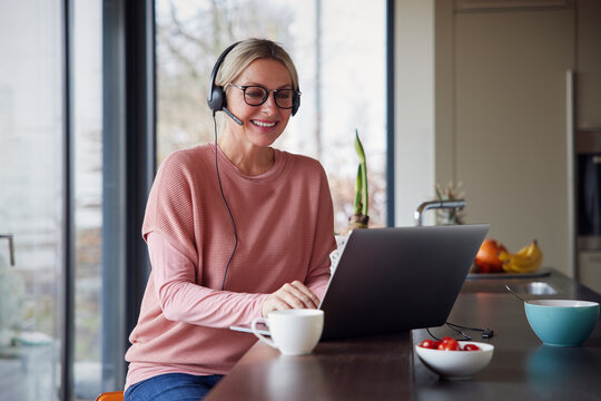 Happy Blond Woman Wearing Headphones Using Laptop In Kitchen