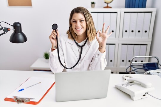 Young hispanic woman wearing doctor uniform holding stethoscope at medical clinic doing ok sign with fingers, smiling friendly gesturing excellent symbol