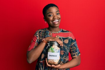 Young african american woman holding savings jar with south african rands money winking looking at...