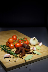 Vegetables and spices on a wooden board on a dark background. Cherry tomatoes and sun-dried tomatoes, garlic, rosemary sprig, bay leaves, black pepper and coarse sea salt.