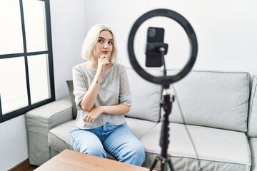 Fototapeta na wymiar Young caucasian woman recording vlog tutorial with smartphone at home looking confident at the camera smiling with crossed arms and hand raised on chin. thinking positive.