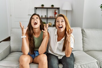 Mother and daughter together sitting on the sofa at home crazy and mad shouting and yelling with aggressive expression and arms raised. frustration concept.