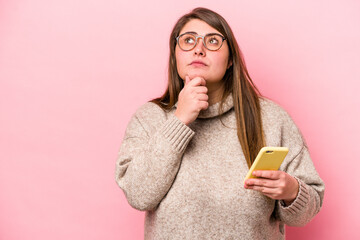 Young caucasian overweight woman holding a mobile phone isolated on pink background looking sideways with doubtful and skeptical expression. - obrazy, fototapety, plakaty