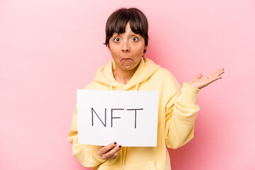 Young hispanic woman holding a NFT placard isolated on pink background shrugs shoulders and open eyes confused.