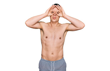 Fototapeta premium Handsome young man wearing swimwear shirtless doing ok gesture like binoculars sticking tongue out, eyes looking through fingers. crazy expression.