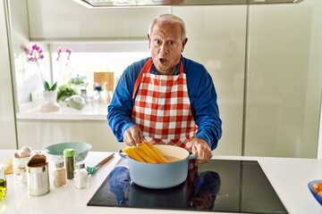 Senior man with grey hair cooking spaghetti at home kitchen scared and amazed with open mouth for...