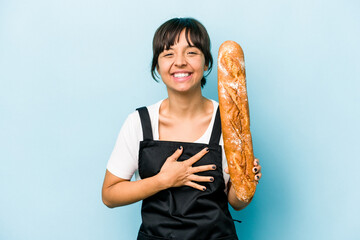 Young hispanic baker woman holding a loaf of bread isolated on blue background laughing and having...