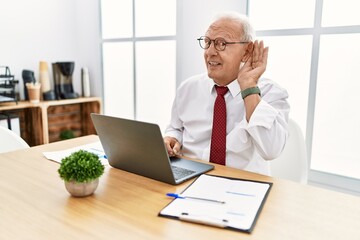 Senior man working at the office using computer laptop smiling with hand over ear listening an hearing to rumor or gossip. deafness concept.