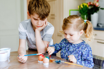 Obraz premium Excited little toddler girl and older brother, school kid boy coloring eggs for Easter. Two cute children, siblings looking surprised at colorful eggs, celebrating holiday with family.