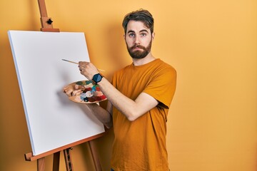 Caucasian man with beard standing drawing with palette by painter easel stand depressed and worry...