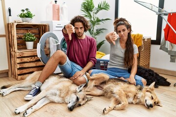 Young hispanic couple doing laundry with dogs looking unhappy and angry showing rejection and negative with thumbs down gesture. bad expression.