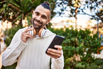 Fototapeta premium Handsome middle age man with beard standing happy and confident outdoors using smartphone and wearing headphones