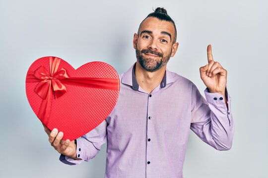 Young hispanic man holding valentine gift smiling with an idea or question pointing finger with happy face, number one