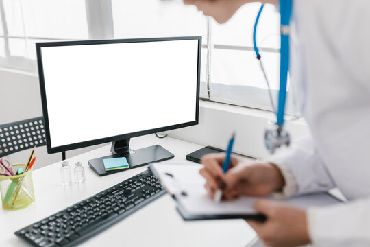 Doctor Writing On Clipboard While Looking At Computer Screen At Clinic