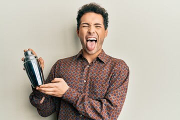 Young handsome man preparing cocktail mixing drink with shaker sticking tongue out happy with funny expression.