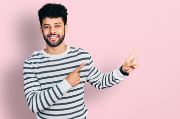 Young arab man with beard wearing casual striped sweater smiling and looking at the camera pointing with two hands and fingers to the side.