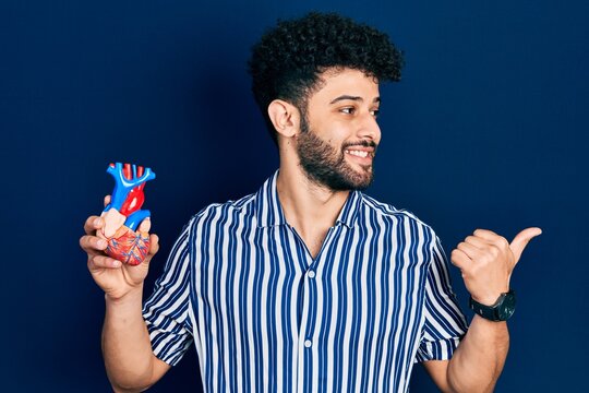 Young arab man with beard holding heart organ pointing thumb up to the side smiling happy with open mouth
