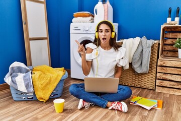 Young hispanic woman studying while waiting for laundry surprised pointing with finger to the side, open mouth amazed expression.