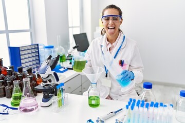 Fototapeta premium Young hispanic woman working at scientist laboratory sticking tongue out happy with funny expression.