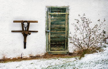 Part of plough, hanging on white wall of farmhouse, green scratched door and shrub on snowy yard.