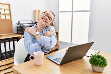 Young redhead woman working at the office using computer laptop hugging oneself happy and positive, smiling confident. self love and self care