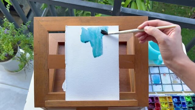POV female artist painting blue background on cotton watercolour paper pad on wooden easel outdoors in summer.First person view of painter making art.