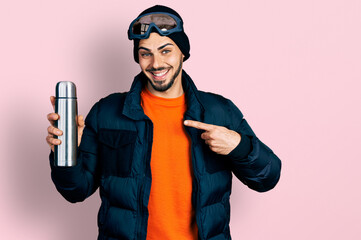 Young hispanic man with beard wearing snow wear and sky glasses holding thermo smiling happy pointing with hand and finger