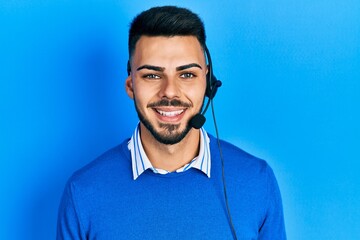 Young hispanic man with beard wearing operator headset at the call center office looking positive...