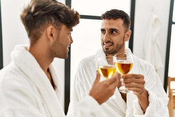 Two hispanic men couple toasting with champagne sitting on massage table at beauty center