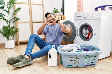Young hispanic man putting dirty laundry into washing machine smiling in love doing heart symbol shape with hands. romantic concept.