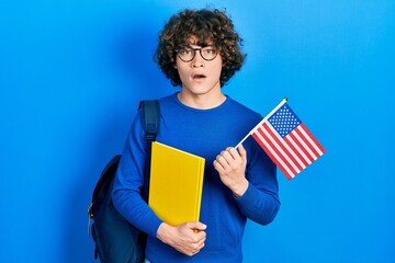 Handsome young man exchange student holding usa flag in shock face, looking skeptical and...