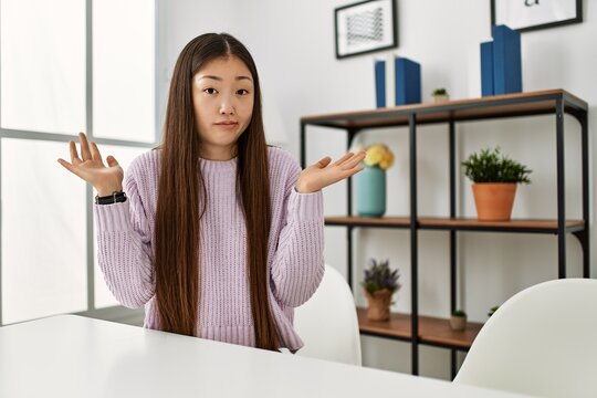 Young chinese girl wearing casual clothes sitting on the table at home clueless and confused expression with arms and hands raised. doubt concept.