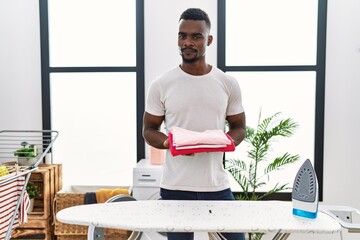 Young african man holding folded laundry after ironing relaxed with serious expression on face....