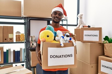 Young african man wearing volunteer t shirt and christmas hat holding donations smiling with a happy and cool smile on face. showing teeth.