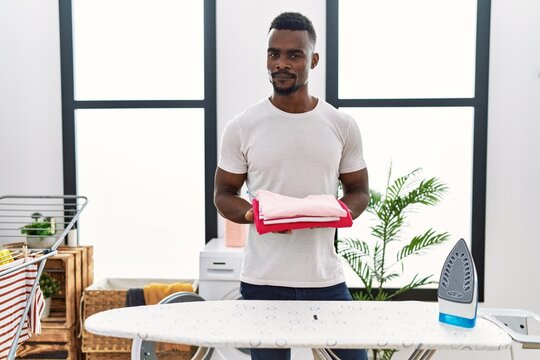 Young african man holding folded laundry after ironing relaxed with serious expression on face. simple and natural looking at the camera.