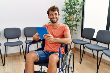 Young hispanic man using touchpad sitting on wheelchair at clinic waiting room.