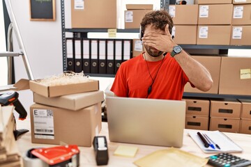 Young hispanic call center agent man working at warehouse covering eyes with hand, looking serious and sad. sightless, hiding and rejection concept