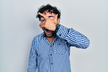 Young hispanic man wearing casual clothes peeking in shock covering face and eyes with hand, looking through fingers with embarrassed expression.