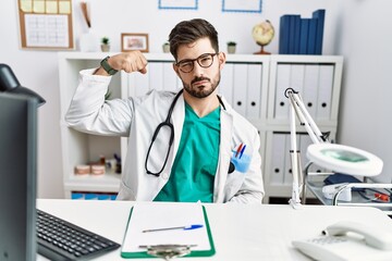 Young man with beard wearing doctor uniform and stethoscope at the clinic strong person showing arm muscle, confident and proud of power