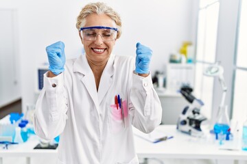 Fototapeta premium Middle age blonde woman working at scientist laboratory excited for success with arms raised and eyes closed celebrating victory smiling. winner concept.